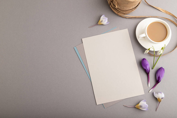Gray paper sheet mockup with spring snowdrop crocus and galanthus flowers and cup of coffee on gray background. Blank, business card, top view, flat lay, copy space, still life. spring concept. - Photo, Image