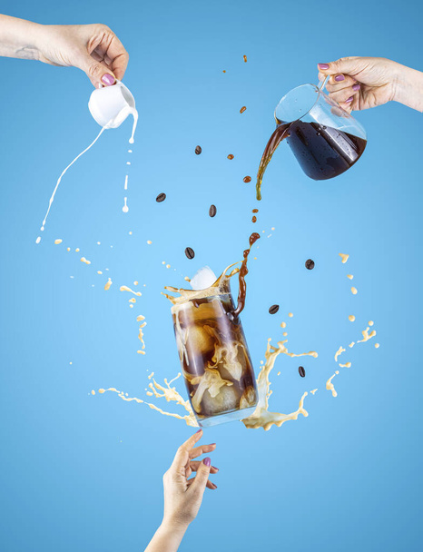 Woman hand support fly glass of tasty brew cold coffee and ice with splash, women hands is pouring homemade sour cream from small jar and coffee from kettle to glassful, ingredients falling - Photo, Image