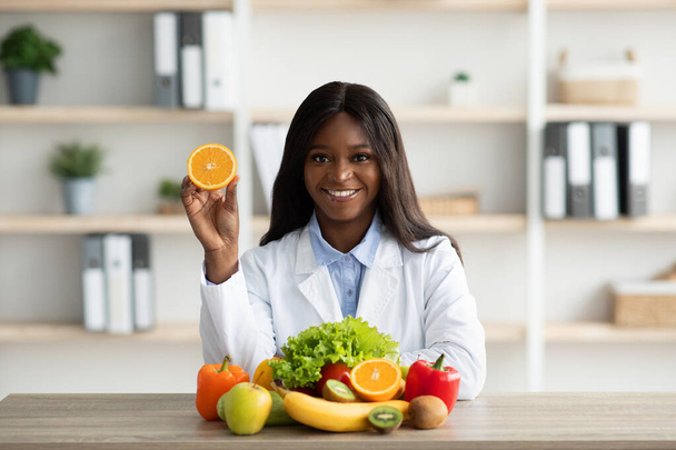 Beginnen Sie Ihren Tag mit frischen Früchten. Glückliche schwarze Ernährungsberaterin mit orangefarbener Hälfte im Büro sitzend und lächelnd - Foto, Bild