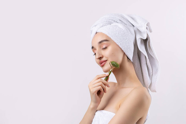Beautiful girl with thick eyebrows and perfect skin at white background, towel on head, beauty photo. Holds a cosmetic roller. - Valokuva, kuva