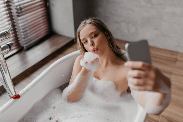 Jolie dame couchée dans la baignoire et prenant selfie sur smartphone, se reposant dans la salle de bain et jouant avec de la mousse - Photo, image