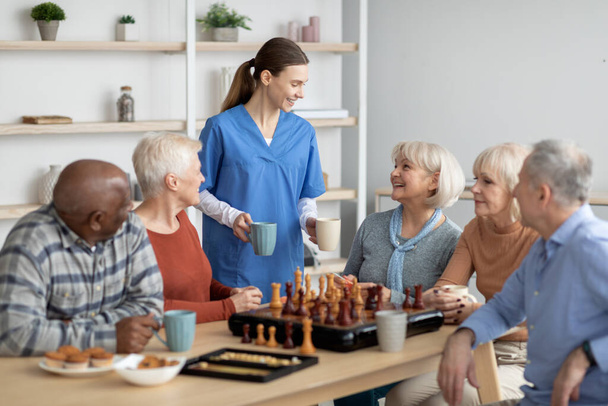 Friendly nurse holding mugs, talking to elderly people, serving tea - Foto, afbeelding