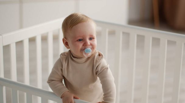 Beruhigungsmittel für Kleinkinder. Kleines Baby mit Schnuller im Kinderbett stehend, Blick seitlich auf Schlafzimmer, Panorama, freier Raum - Foto, Bild