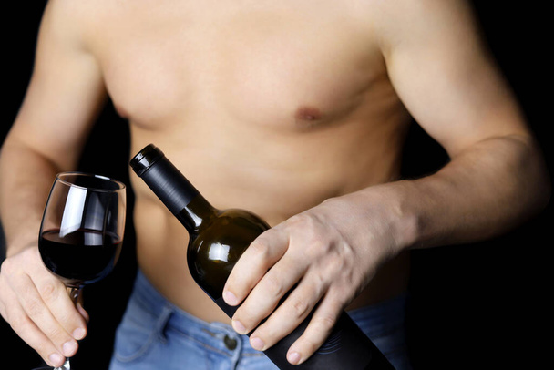 Muscular man in with naked torso pouring red wine in a glass from the bottle. Concept of alcohol party, romantic date - Фото, изображение