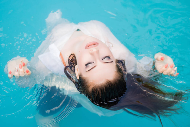 Top view, portrait of a young, beautiful woman floating in the pool, wearing a white shirt. Rest in the water in hot weather. - Foto, immagini