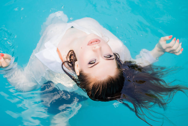 Top view, portrait of a young, beautiful woman floating in the pool, wearing a white shirt. Rest in the water in hot weather. - Zdjęcie, obraz