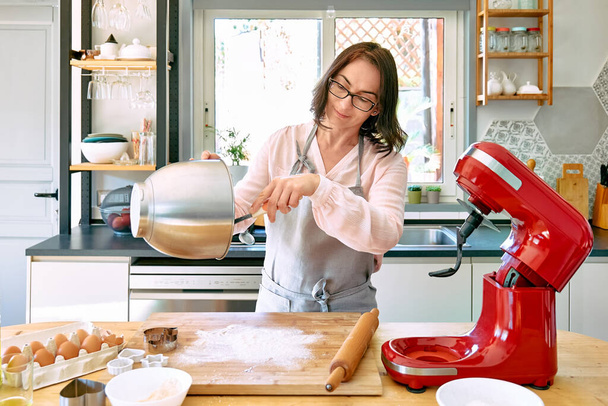 Femme portant tablier pâtisserie biscuits dans une cuisine confortable. La femme au foyer prend la pâte dans le bol du mélangeur planétaire. Gâteaux ou tartes faits maison. - Photo, image