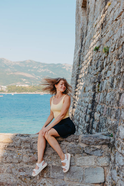 Beautiful girl sitting on a stone wall, in background is the blue sea, Budva, Montenegro. - Photo, image