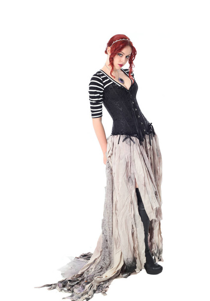 full length portrait of girl with red hair wearing black corset , torn gothic fantasy gown and leather boots. standing pose, isolated on white studio background. - Zdjęcie, obraz