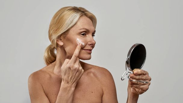 Woman looking at mirror and smearing cream on face - Photo, Image