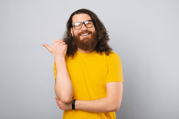 Aufgeregt zeigt ein bärtiger Mann im gelben T-Shirt mit langen Haaren mit dem Daumen zur Seite. Studioaufnahme vor grauem Hintergrund. - Foto, Bild