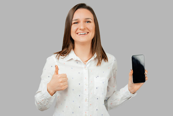 Nahaufnahme Porträt einer jungen Frau in weißem Hemd, die ein leeres Bildschirmtelefon und den Daumen nach oben zeigt. Studioaufnahme vor grauem Hintergrund. - Foto, Bild