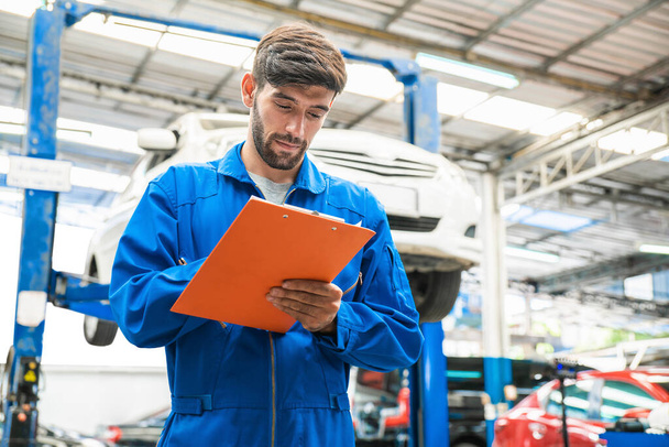 A kék munkaruhás szerelő egyenruhában ellenőrzi a jármű karbantartási ellenőrzőlistáját homályos felemelt autóval a háttérben. Gépjármű-javítási szolgáltatás, Szakmai foglalkozás. - Fotó, kép