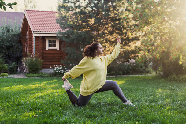 Caucasian over-weighted woman in sports clothes doing physical exercises on backyard on green grass with wooden country house and high trees in background. Body positive - Foto, afbeelding