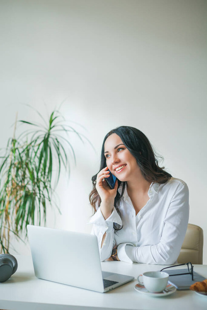 Die junge Geschäftsfrau arbeitet am Computer und telefoniert. Eine schöne dunkelhaarige Frau in weißem Hemd arbeitet online in einem hellen Büro. Kaffee trinken und Croissant essen. - Foto, Bild