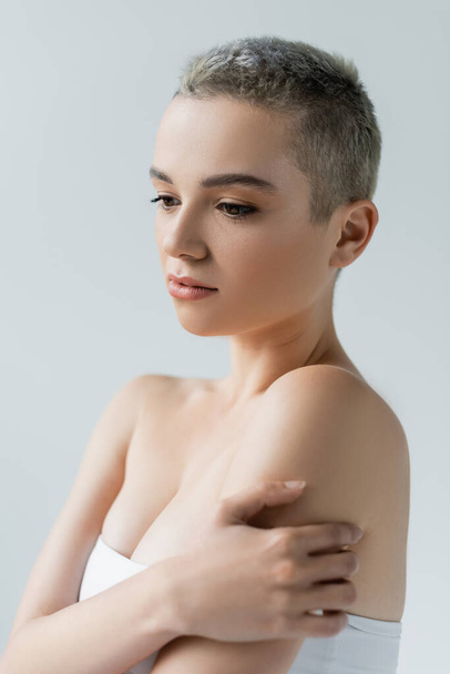 junge Frau mit kurzen Haaren und nackten Schultern umarmt sich vereinzelt auf grau - Foto, Bild