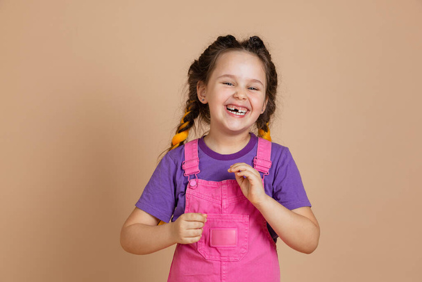 Porträt einer lachenden kleinen Frau mit Kanekalon-Zöpfen und fehlenden Zähnen, die in die Kamera blickt und einen rosafarbenen Overall und ein lila T-Shirt auf beigem Hintergrund trägt - Foto, Bild