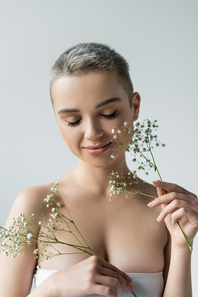 pretty woman with naked shoulders and closed eyes smiling near branches of gypsophila isolated on grey - Zdjęcie, obraz