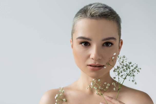 charming woman with naked shoulders looking at camera near small white flowers isolated on grey - Foto, Imagem