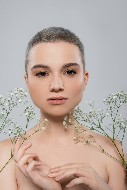portrait of young woman with natural makeup looking at camera near white tiny flowers isolated on grey - Foto, Imagen