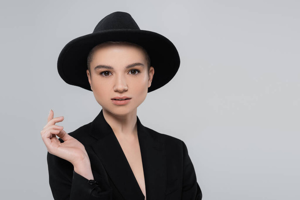 stylish woman in black fedora hat and blazer looking at camera isolated on grey - 写真・画像