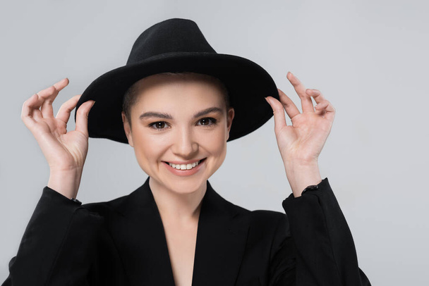 Vorderansicht einer lächelnden Frau, die ihren schwarzen Hut mit Krempe isoliert auf grau einstellt - Foto, Bild