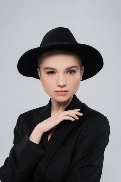 charming woman in black blazer and brim hat looking at camera isolated on grey - Photo, Image