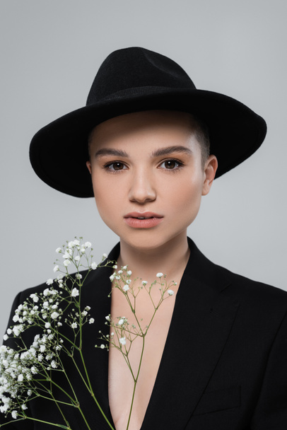 woman in black brim hat, with natural makeup, looking at camera near tiny flowers isolated on grey - Фото, изображение