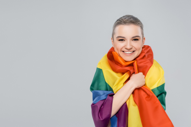 joyful woman looking at camera while wrapping in lgbt flag isolated on grey - Foto, imagen