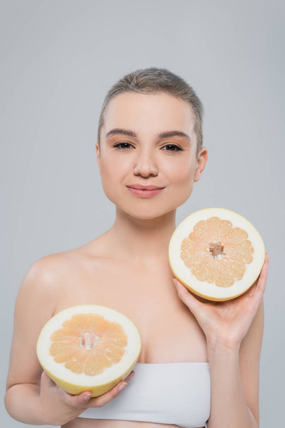 pleased woman with perfect skin holding halves of juicy grapefruit isolated on grey - Фото, зображення