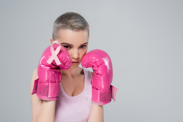 junge Frau mit pinkfarbener Schleife auf Boxhandschuh in Verteidigungsposition isoliert auf grau - Foto, Bild