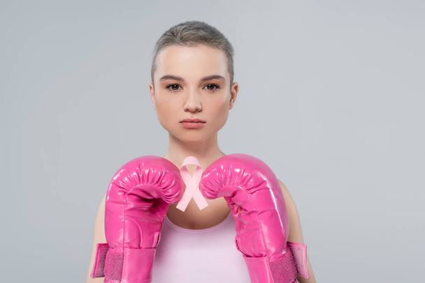 mujer con el pelo corto, en guantes de boxeo de color rosa, la celebración de cáncer de mama cinta de conciencia aislado en gris - Foto, Imagen