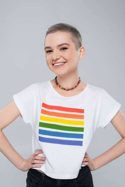 cheerful young woman in lgbt colors t-shirt standing with hands on hips isolated on grey - Photo, Image