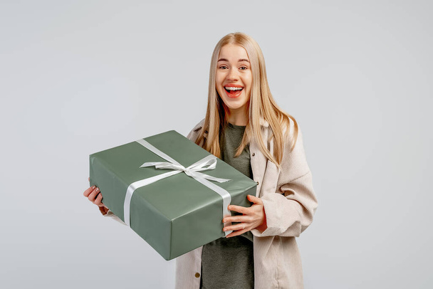 Portrait of Happy young blonde woman in trendy shirt holding big green gift box and looking at the camera over gray background - Photo, Image