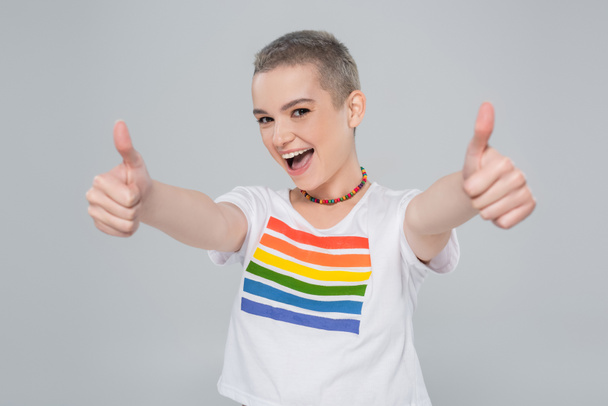 vrolijke vrouw in regenboog kleuren t-shirt tonen duimen omhoog geïsoleerd op grijs - Foto, afbeelding