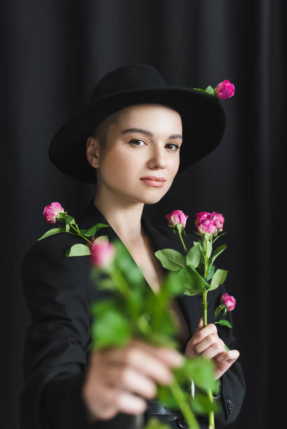 pretty woman in brim hat and black blazer holding pink roses on dark background - Photo, Image