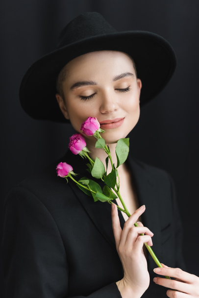 mujer joven con los ojos cerrados sosteniendo pequeñas rosas rosadas cerca de los labios en negro  - Foto, imagen