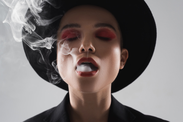front view of stylish woman with closed eyes exhaling smoke isolated on grey - 写真・画像