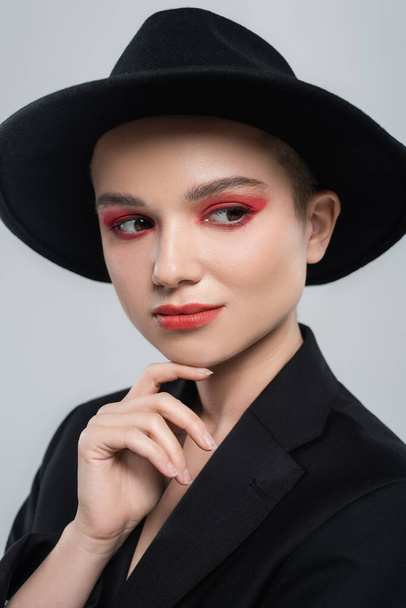 young woman with carmine red makeup, in black brim hat, touching chin isolated on grey - Foto, imagen