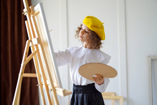 Portrait of little girl artist in french hat who paints picture in studio while holds pallete  - Фото, изображение