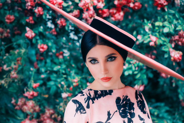 Fantasy retro woman style. lady in pink hat wide brimmed. Natural cosmetics concept, cat eyes makeup, cute face red lipstick lips. Spring blooming garden green rose bushes. Portrait girl fashion  - Foto, immagini