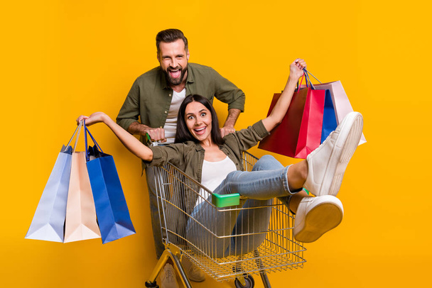 Photo of young cheerful couple have fun shop discount holiday present drive trolley store isolated over yellow color background - 写真・画像