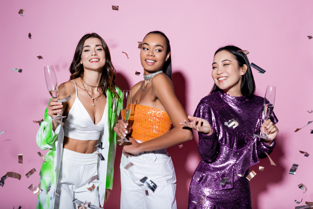 cheerful multiethnic women in trendy outfits holding glasses of champagne near falling confetti on pink  - Foto, immagini