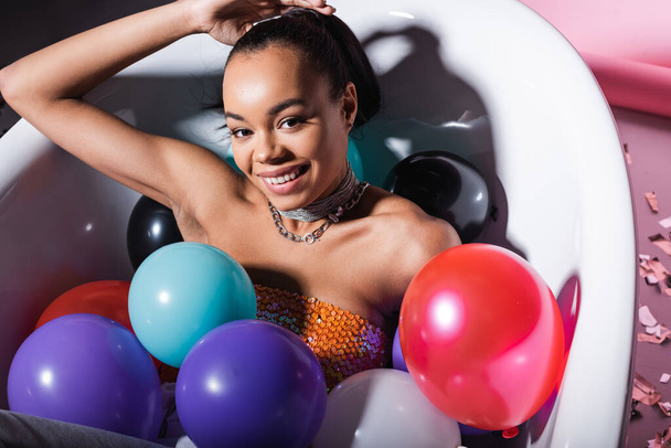 high angle view of pleased african american woman lying in bathtub with colorful balloons  - Foto, Bild