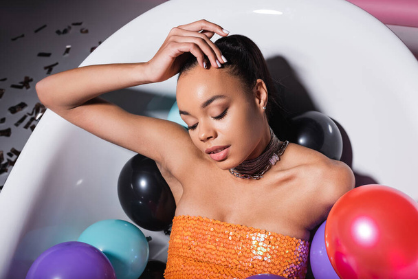 high angle view of young african american model lying in bathtub with colorful balloons  - Fotografie, Obrázek