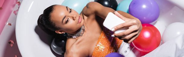 high angle view of african american model lying in bathtub with colorful balloons and taking selfie on smartphone, banner - Foto, imagen