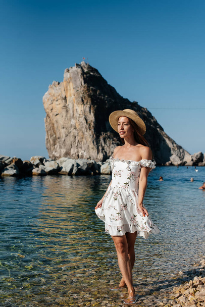 A beautiful young woman in a hat, glasses and a light dress is walking along the ocean shore against the background of huge rocks on a sunny day. Tourism and tourist trips. - Foto, Imagen