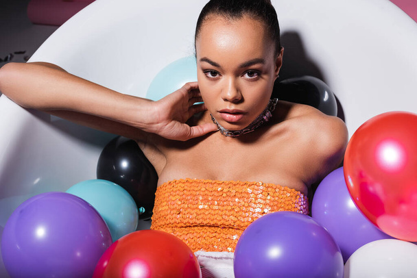 happy african american woman lying in bathtub with colorful balloons looking at camera - Foto, imagen