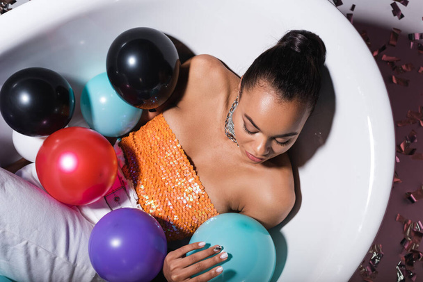 top view of brunette african american woman lying in bathtub with balloons  - Photo, Image