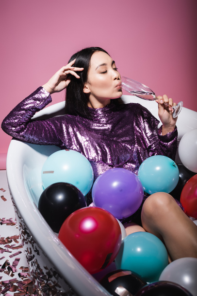 asian woman in purple dress lying in bathtub with colorful balloons and drinking champagne on pink  - Foto, Imagen